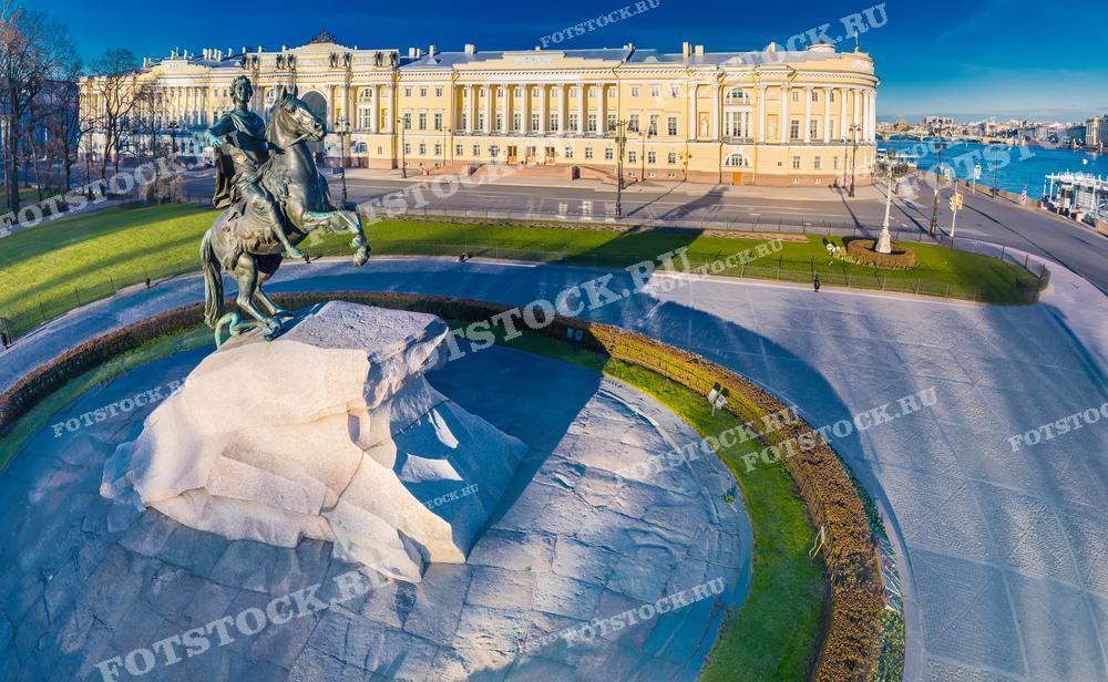 Фотография показывает памятник Петру Великому, известный как Медный всадник, расположенный на Сенатской площади в Санкт-Петербурге. На заднем плане изображены фасады зданий, в том числе исторического здания Сената. Светлое небо и яркое солнце добавляют фото ясности и величия, подчёркивая значимость памятника и архитектурной среды. Это изображение идеально подходит для исторических и культурных публикаций, туристических проспектов и учебных материалов о Санкт-Петербурге.